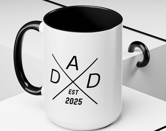 May include: White ceramic mug with a black interior, handle, and rim. The mug features the word "DAD" in a crossed-out design with "EST 2025" printed below in black. The mug is on a white surface.