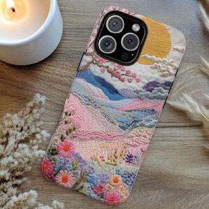 May include: A phone case featuring an embroidered landscape design with a sun, mountains, and flowers in shades of pink, blue, and yellow. The case has a black camera module and a white border.