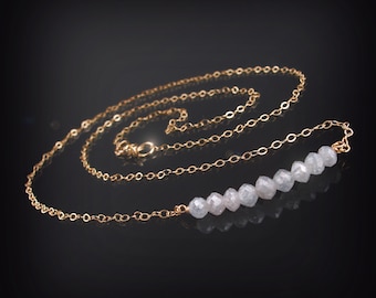 May include: A gold chain necklace with a row of white faceted beads. The necklace is on a black background.