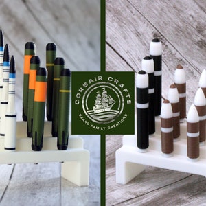May include: Two sets of miniature model rockets on white display stands. The rockets are painted in various colors, including black, white, green, orange, and brown. The stands are made of white plastic and have a simple design. The text "CORSAIR CRAFTS Beard Family Creations" is visible on a green label.