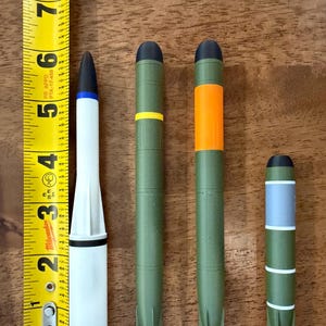 May include: Four cylindrical projectile models arranged vertically next to a yellow Milwaukee measuring tape. The first is white with a black tip and bands. The next two are olive green with black tips and colored bands. The last is shorter, olive green with a grey section and orange fins.