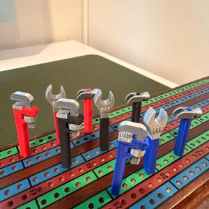May include: A collection of colorful, 3D-printed wrenches and hammers arranged on a wooden cribbage board. The tools are in various colors, including red, black, blue, and silver. The cribbage board has a multi-colored design with numbers.