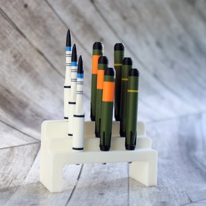 May include: A white plastic stand holds a collection of model missiles. The missiles are painted in various colors, including white, black, blue, green, orange, and yellow.