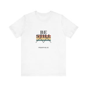 May include: A white short-sleeve t-shirt featuring a graphic design on the chest. The design includes the black text BE STILL above horizontal stripes in orange, yellow, and teal, with a black mountain outline and the text PSALM 46:10 below.