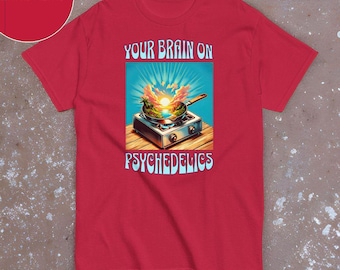 May include: Red t-shirt with a graphic design featuring a frying pan on a stove with a sunset scene inside. The text "YOUR BRAIN ON PSYCHEDELICS" is above and below the image. The shirt has short sleeves and a crew neck.