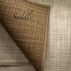 May include: A piece of fabric with a woven texture, featuring a folded corner that reveals a dark brown colour on the top side and a light beige colour on the underside. The word Saddle is printed in black script on the brown section of the fabric.