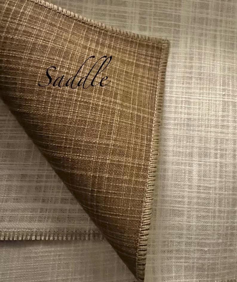 May include: A piece of fabric with a woven texture, featuring a folded corner that reveals a dark brown colour on the top side and a light beige colour on the underside. The word Saddle is printed in black script on the brown section of the fabric.