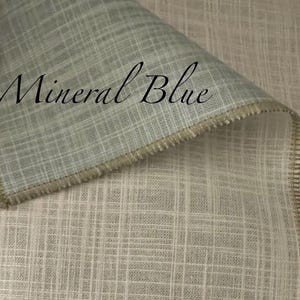 May include: A piece of fabric with a visible grid-like weave pattern in a light mineral blue colour. The textile features a folded edge with a serged seam finish. The text Mineral Blue is printed in a black script font across the fabric surface.