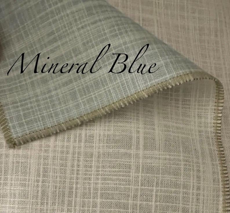 May include: A piece of fabric with a visible grid-like weave pattern in a light mineral blue colour. The textile features a folded edge with a serged seam finish. The text Mineral Blue is printed in a black script font across the fabric surface.