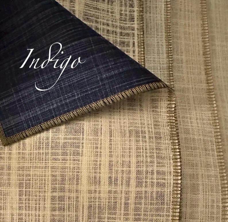 May include: A piece of fabric with a folded corner revealing a dark navy blue side and a light beige side. Both sides feature a visible grid-like weave texture. The word Indigo is written in white cursive script across the dark blue section.