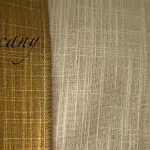 May include: A side-by-side view of two textured fabrics. On the left, a mustard-gold fabric with a visible grid pattern and the word Tuscany written in black script. On the right, a cream-coloured fabric with a vertical slubbed weave texture.