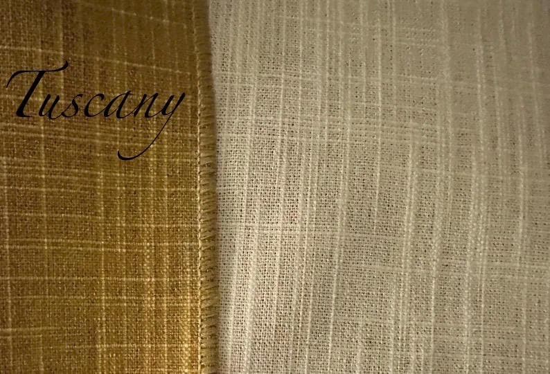 May include: A side-by-side view of two textured fabrics. On the left, a mustard-gold fabric with a visible grid pattern and the word Tuscany written in black script. On the right, a cream-coloured fabric with a vertical slubbed weave texture.