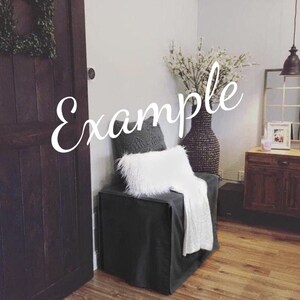 May include: A gray upholstered chair with a white throw blanket and a gray knitted pillow. The chair is in front of a white wall and a dark wooden door. A vase with white flowers is on the right side of the chair.