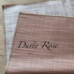 May include: Two rectangular fabric placemats with a visible woven texture. The top placemat is dusty rose coloured with the text "Dusty Rose" printed in black script. The bottom placemat is a light beige colour. Both have finished stitched edges.