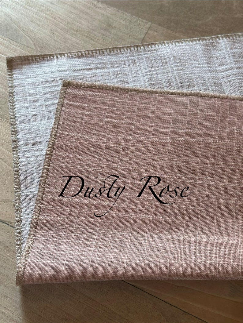 May include: Two rectangular fabric placemats with a visible woven texture. The top placemat is dusty rose coloured with the text "Dusty Rose" printed in black script. The bottom placemat is a light beige colour. Both have finished stitched edges.