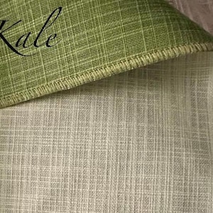 May include: A piece of fabric with a woven grid texture, featuring a folded corner that reveals a sage green top side and a cream-coloured underside. The word Kale is printed in black script on the green section, and the edges are finished.