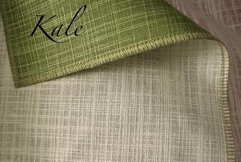 May include: A piece of fabric with a woven grid texture, featuring a folded corner that reveals a sage green top side and a cream-coloured underside. The word Kale is printed in black script on the green section, and the edges are finished.