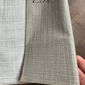 May include: A piece of fabric with a woven texture featuring a light grey side and a tan side labelled with the text Ecru. The material has a visible crosshatch pattern throughout, with a finished stitched edge separating the two distinct colour sections.