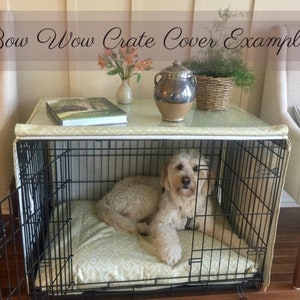 May include: A black metal dog crate with a beige and white patterned cover. The crate is sitting on a table with a book, a vase of flowers, and a ceramic jar. A small dog is lying inside the crate.