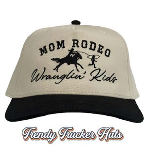 May include: A beige trucker hat with a black brim. The front of the hat features black embroidered text that reads "Mom Rodeo Wranglin' Kids" with a graphic of a person riding a horse and another person roping a calf.