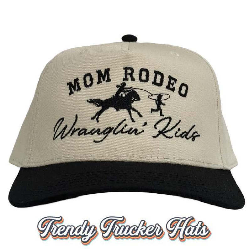 May include: A beige trucker hat with a black brim. The front of the hat features black embroidered text that reads "Mom Rodeo Wranglin' Kids" with a graphic of a person riding a horse and another person roping a calf.