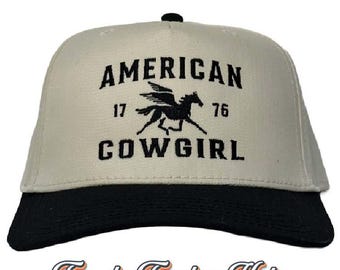 May include: A beige trucker hat with a black bill. The front of the hat features black embroidered text that reads "AMERICAN 17 76 COWGIRL" with a black winged horse graphic.