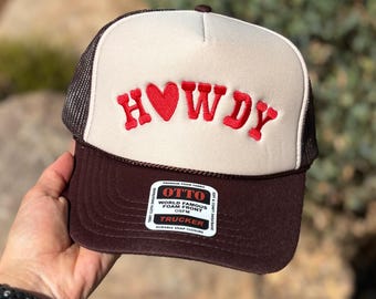 Op de afbeelding: Truckerpet met een wit voorpaneel met het woord "HOWDY" in rode borduursels. De pet heeft een bruine rand en een mesh achterkant. Het label luidt "OTTO".
