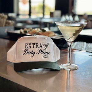May include: White baseball cap with a black brim and the text "EXTRA Dirty Please" embroidered on the front. A martini glass with olives is in the background.