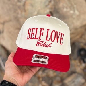 Pode incluir: Boné de beisebol creme e vermelho com a inscrição bordada "SELF LOVE Club" em vermelho. O boné tem uma aba vermelha e um botão vermelho. O boné tem um ajuste clássico com um perfil médio.