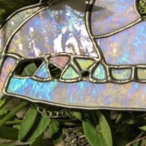 May include: A stained glass animal skull with iridescent glass pieces in shades of blue, purple, and green. The skull is set against a background of green foliage.