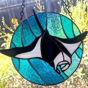 Puede incluir: Una ventana de vitral colgada de una cadena negra. La ventana es redonda y presenta una manta raya nadando en agua turquesa y verde.