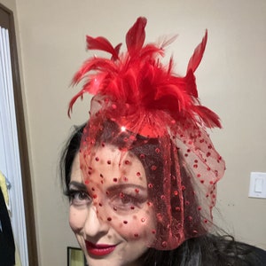 May include: A red feather hat with a red tulle veil and red polka dots. The hat is worn on a person's head.