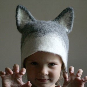 May include: A child wearing a gray and white wolf hat with large ears. The child is making a claw-like gesture with both hands.