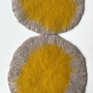 以下が含まれることがあります： グレーの外側の輪と黄色のセンターを持つ、2つの丸いフェルトコースター。コースターはウール製で、柔らかく質感のある表面をしています。