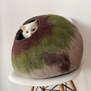 May include: A white cat with black markings peeks out of a green, brown, and beige felt cat bed. The bed is round and sits on a white chair with a wooden base.