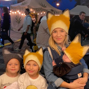 Peut inclure: Une famille de quatre personnes portant des chapeaux sur le thème du renard. La mère porte un bébé dans un porte-bébé. Les deux enfants plus âgés se tiennent à côté d'elle. Les chapeaux sont en feutre et ont de longues oreilles tombantes. Les chapeaux sont de couleurs différentes, mais ils ont tous un design similaire.