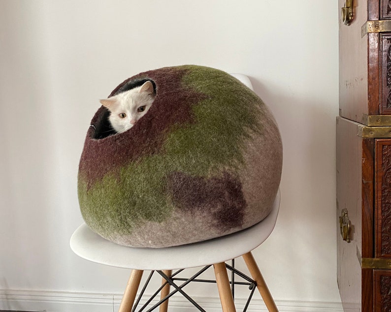 May include: A white cat with black markings peeks out of a green, brown, and beige felt cat bed. The bed is round and sits on a white chair with a wooden base.