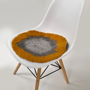 May include: A mustard yellow and gray wool chair cushion with a round shape. The cushion is placed on a white chair with wooden legs.