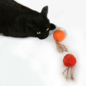 May include: A black cat looking at three orange and red felt balls with strings attached. The balls are on a white surface.