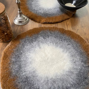 May include: Two round, brown, grey, and white felt placemats with a gradient design. The placemats are on a wooden table.