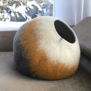 May include: A round, gray, yellow, and white felt cat bed with a black opening. The bed is on a gray couch.