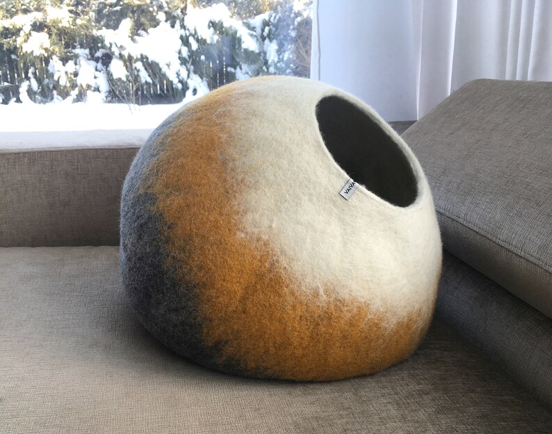 May include: A round, gray, yellow, and white felt cat bed with a black opening. The bed is on a gray couch.