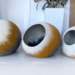 May include: Three felt cat beds in different sizes. The beds are shaped like cocoons and have a white, gray, and yellow ombre design. The largest bed is labeled "Large cocoon", the middle bed is labeled "Medium cave", and the smallest bed is labeled "Small cocoon".