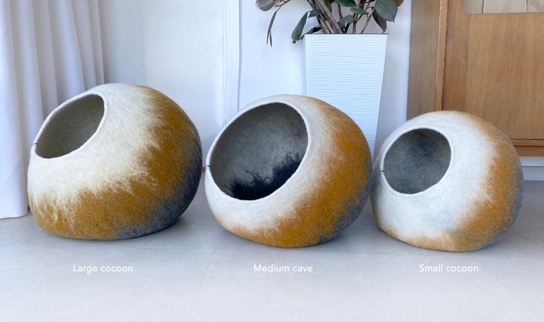 May include: Three felt cat beds in different sizes. The beds are shaped like cocoons and have a white, gray, and yellow ombre design. The largest bed is labeled "Large cocoon", the middle bed is labeled "Medium cave", and the smallest bed is labeled "Small cocoon".