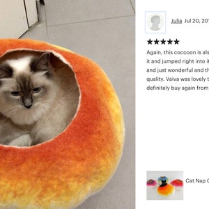 May include: A cat naps inside a bright orange and yellow felt cat bed. The bed is shaped like a cocoon and has a round opening. The bed is labeled "Vaiva Cat".