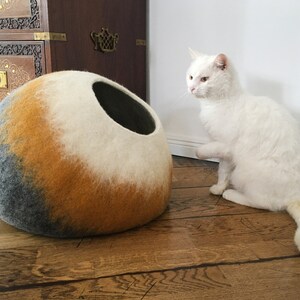 May include: A white cat with a long tail sits on a wooden floor, looking at a round, gray, orange, and white felt cat bed.