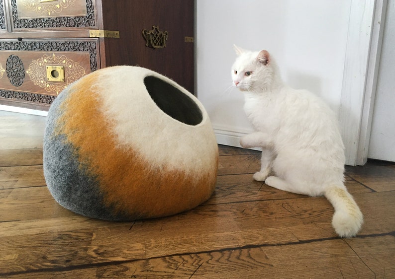 May include: A white cat with a long tail sits on a wooden floor, looking at a round, gray, orange, and white felt cat bed.