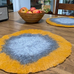 May include: Two round, felted placemats in a light gray color with a yellow border. The placemats are on a wooden table.
