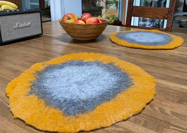May include: Two round, felted placemats in a light gray color with a yellow border. The placemats are on a wooden table.