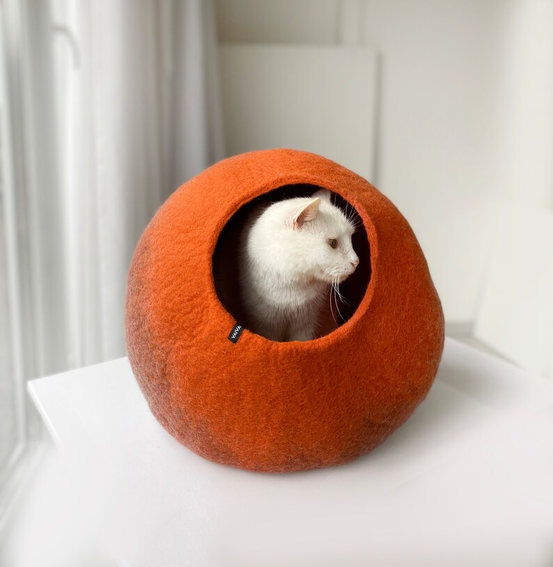 Op de afbeelding: Een witte kat zit in een rond, oranje kattenbed van vilt. Het bed heeft een donkerbruine accent rond de opening.